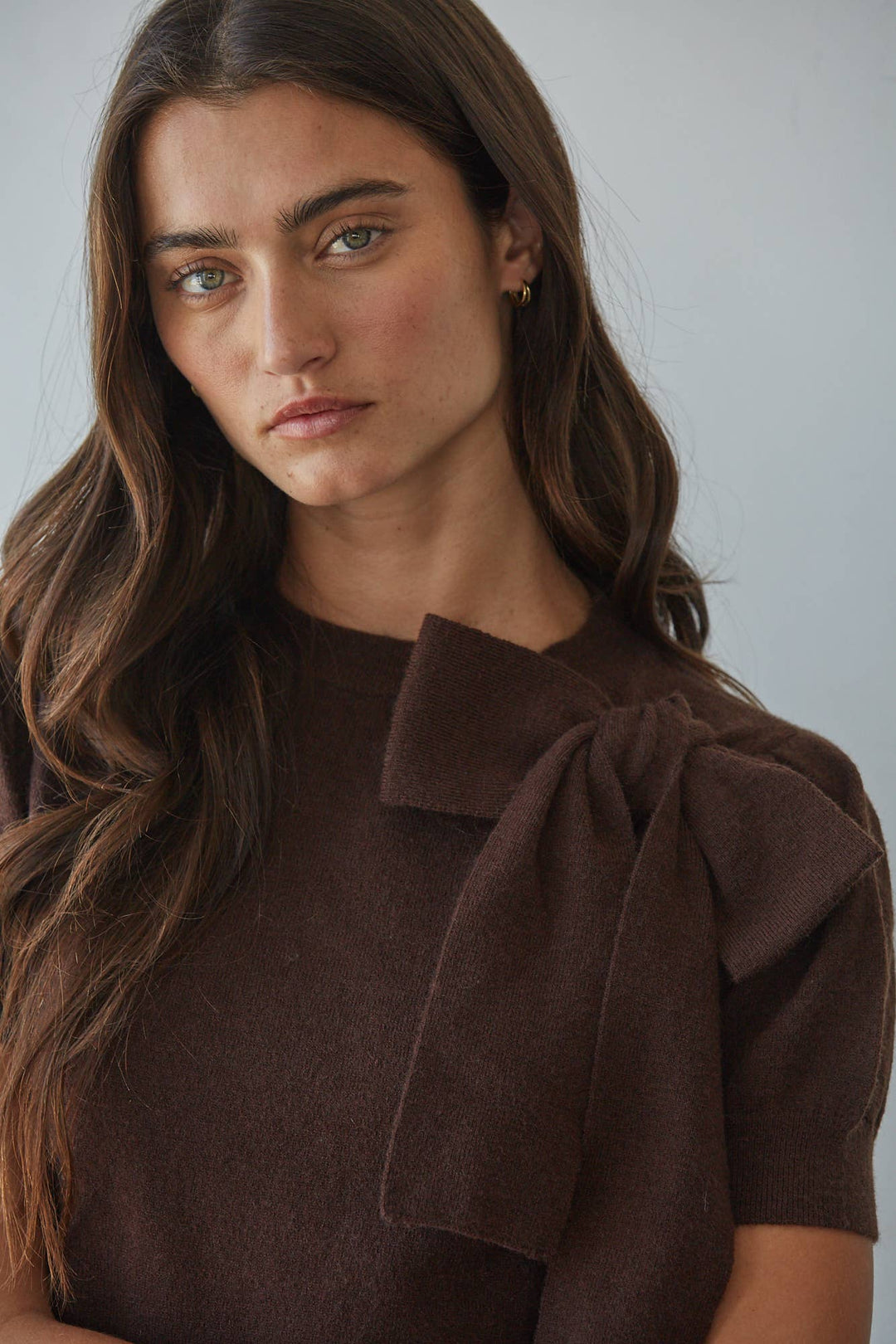 A close-up of a woman wearing a dark brown short-sleeve top with a large bow detail on the shoulder, set against a plain light gray background.