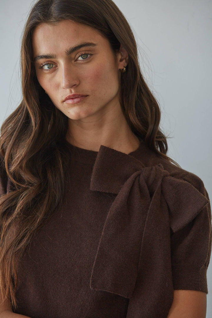 A close-up of a woman wearing a dark brown short-sleeve top with a large bow detail on the shoulder, set against a plain light gray background.