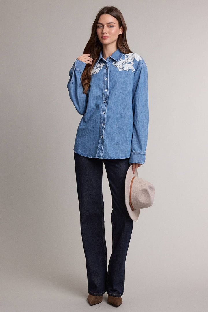 A woman modeling a light blue denim button-up shirt with white floral embroidery on the chest, paired with dark blue jeans and holding a beige wide-brim hat against a plain light gray studio background.