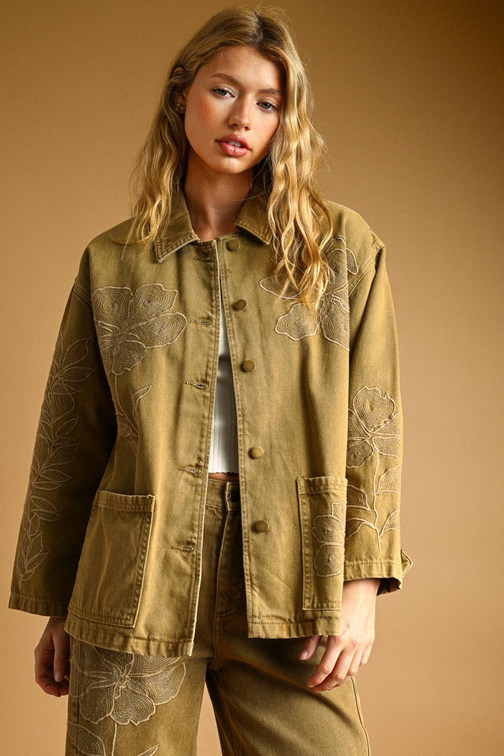 Woman wearing a light brown denim jacket with floral embroidery on a beige background