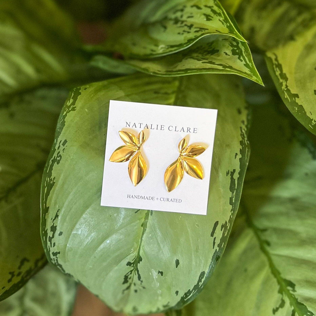 Gold earrings on a card with 'Natalie Clare' branding, placed on a green leafy background.
