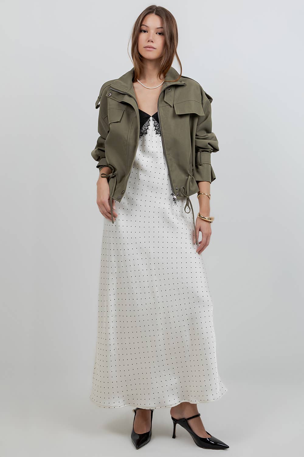 A model stands against a light gray studio background wearing an olive green utility jacket over a white eyelet maxi dress, with black pointed-toe heels.