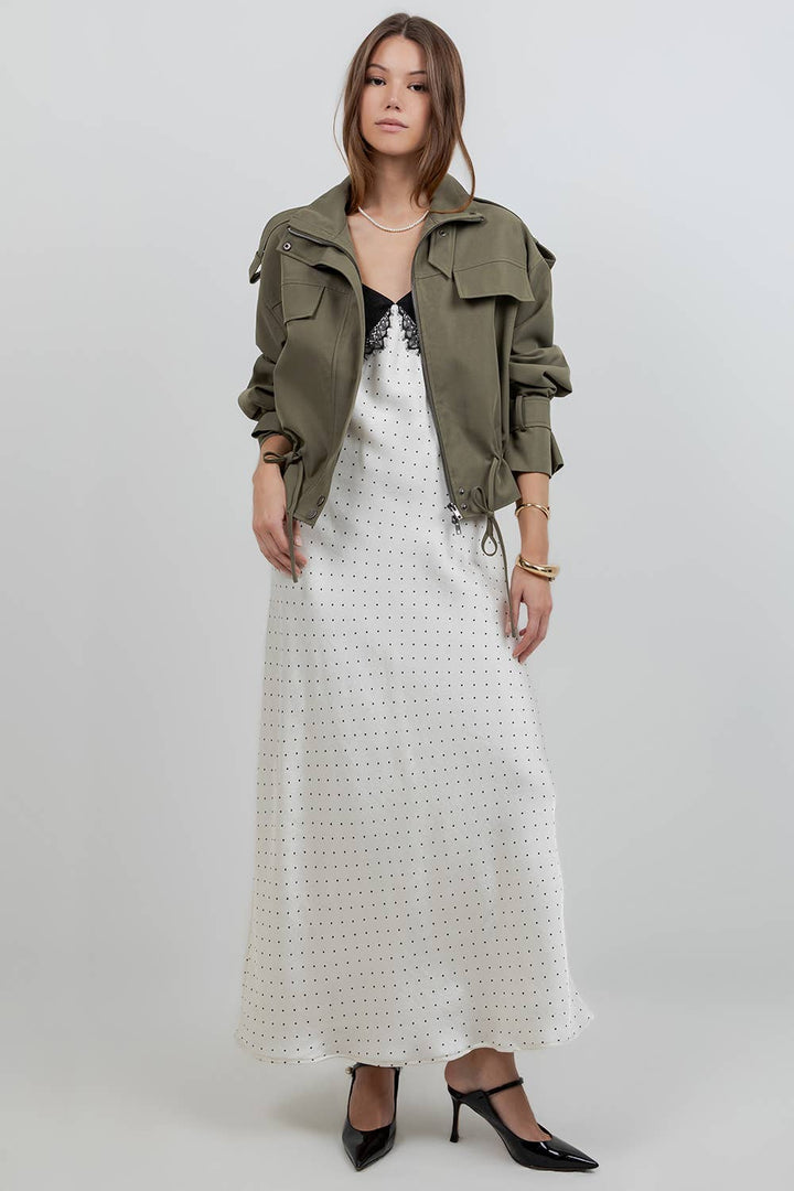 A model stands against a light gray studio background wearing an olive green utility jacket over a white eyelet maxi dress, with black pointed-toe heels.