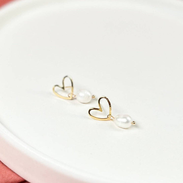 A pair of gold heart-shaped drop earrings with small white pearl beads, displayed on a white ceramic dish.
