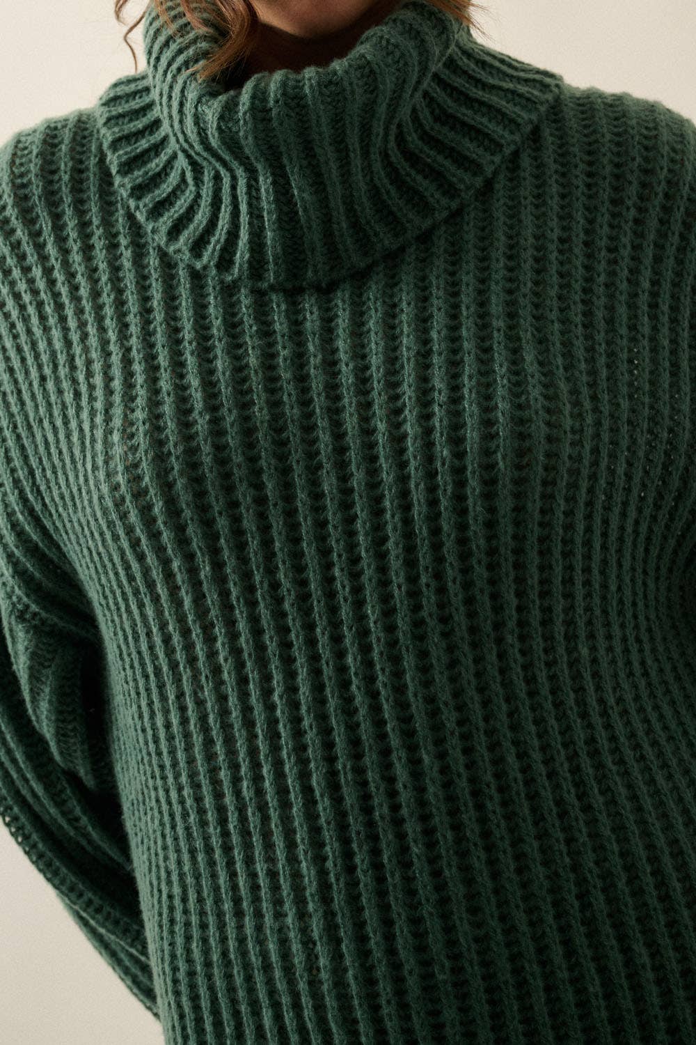 A close-up of a person wearing a dark forest green ribbed knit turtleneck sweater, showing the detailed texture of the knit fabric.