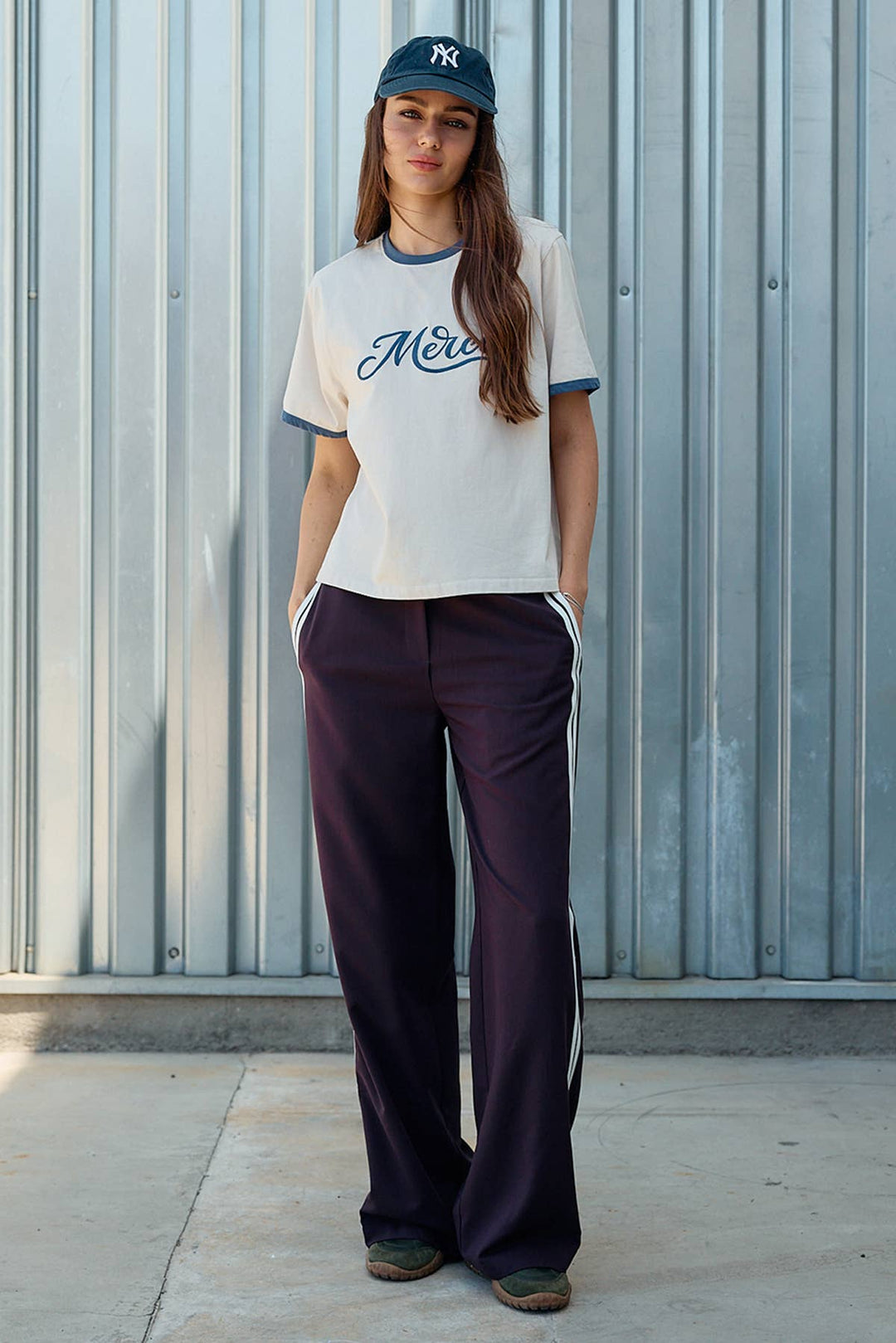 A woman stands in front of a corrugated metal wall wearing a cream ringer T-shirt with navy blue trim and script lettering, dark wide-leg pants with white side stripes, and a navy baseball cap.