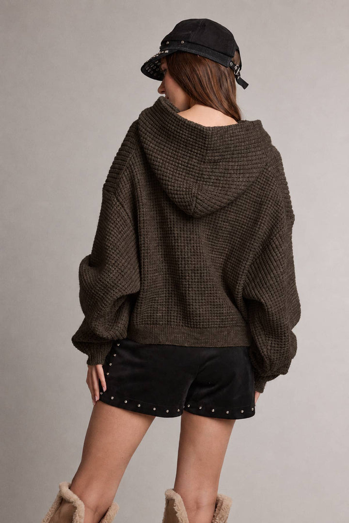 A model shown from the back wearing a dark brown waffle-knit hooded sweater, black studded shorts, a black bucket hat, and beige boots against a neutral gray background.