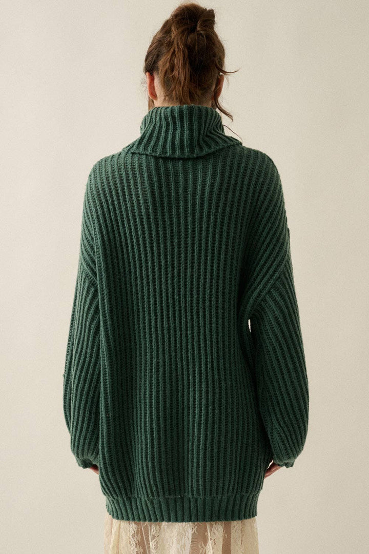 A person shown from the back wearing an oversized forest green ribbed knit turtleneck sweater, paired with a light beige skirt, against a plain light beige background.
