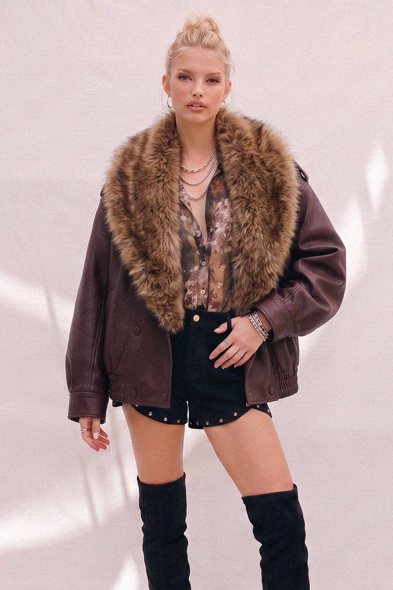 A model wearing a dark brown faux-leather jacket with a large brown fur collar, styled with a patterned blouse, black shorts, and black over-the-knee boots against a light pink wall.