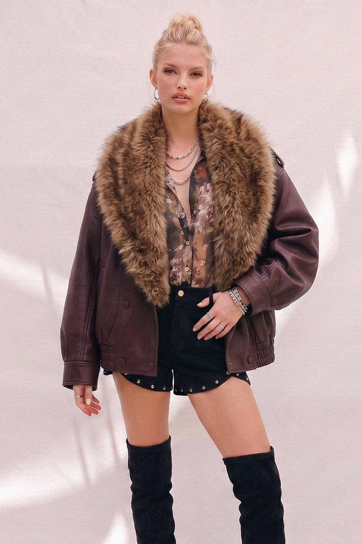 A model wearing a dark brown faux-leather jacket with a large brown fur collar, styled with a patterned blouse, black shorts, and black over-the-knee boots against a light pink wall.