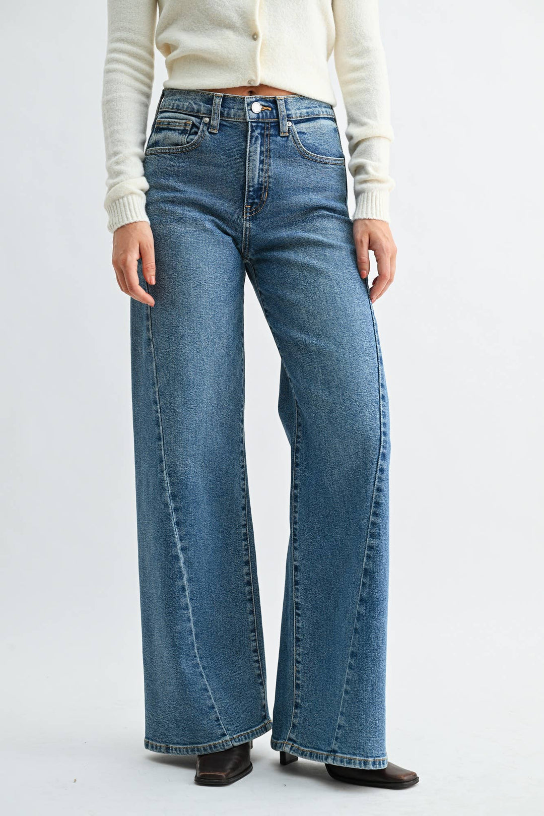 A model wearing a pair of wide-leg blue denim jeans with a medium faded wash, styled with a cream-colored knit cardigan and dark brown flat shoes against a plain white studio background.