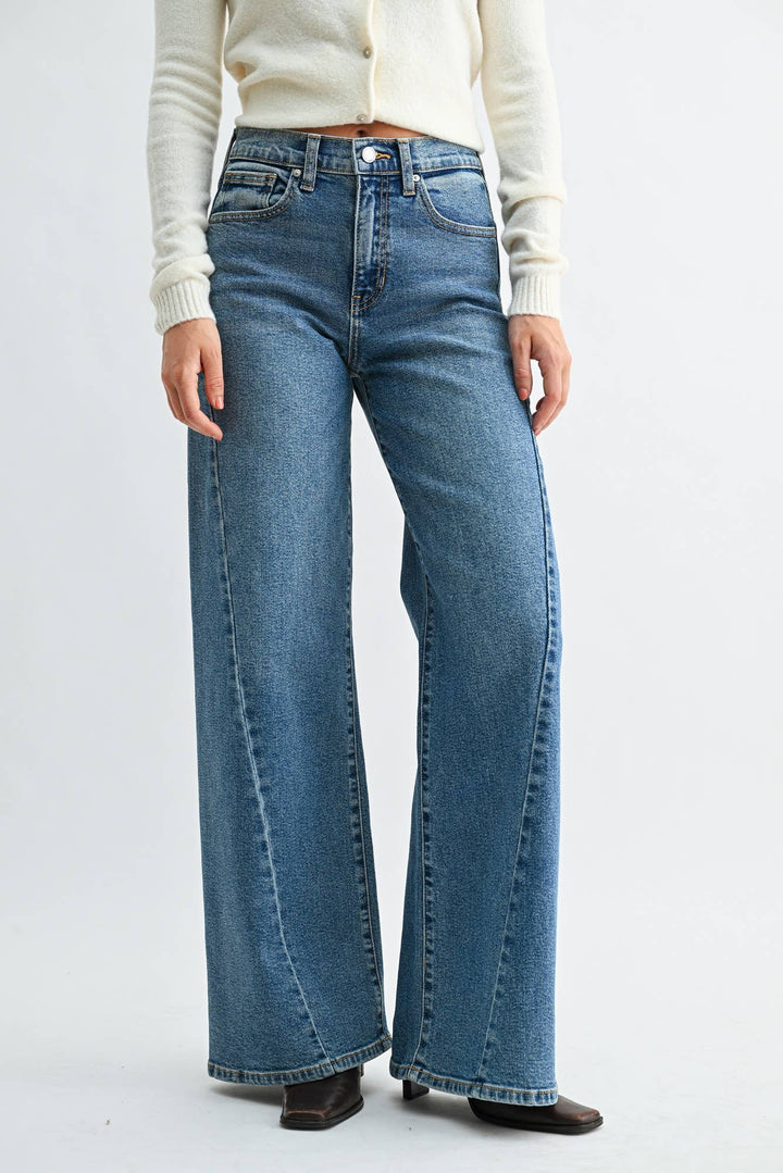A model wearing a pair of wide-leg blue denim jeans with a medium faded wash, styled with a cream-colored knit cardigan and dark brown flat shoes against a plain white studio background.