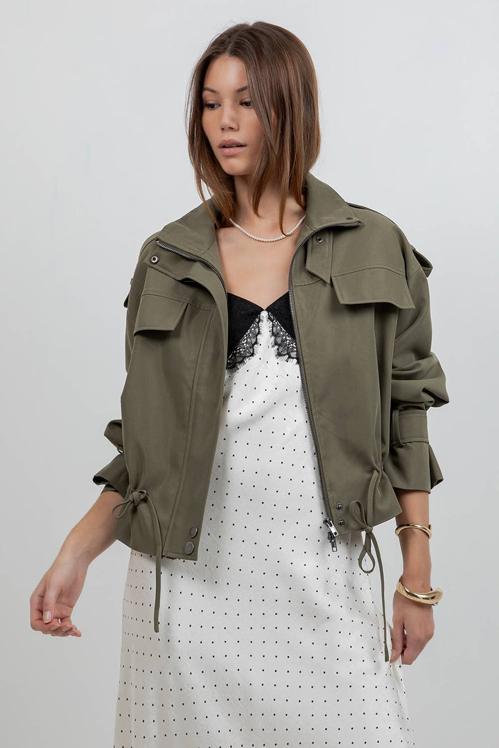 A woman wearing an olive green military-style jacket over a white polka dot dress with a black lace-trimmed top, photographed against a light gray studio background.