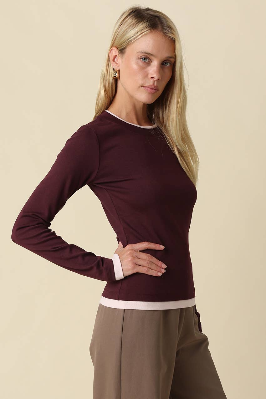 A blonde woman modeling a long-sleeve crewneck long sleeve t-shirt top in deep burgundy with pink contrast trim at the collar, cuffs, and hem, paired with taupe trousers, against a solid beige background.