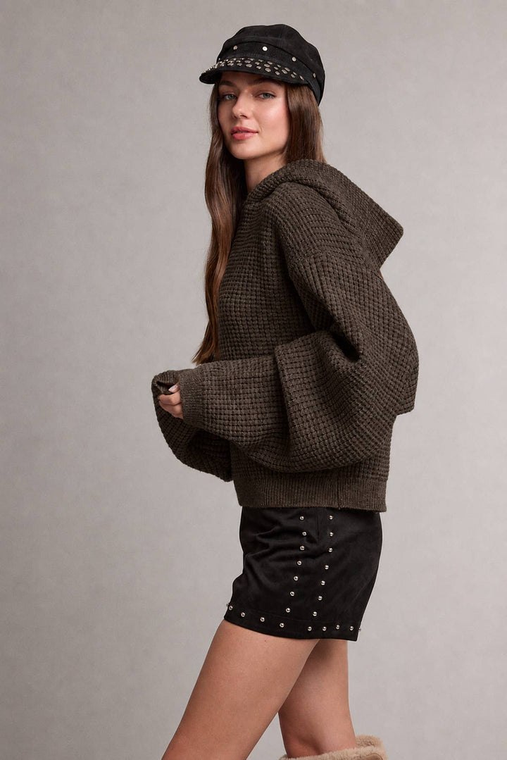 A woman modeling a dark brown waffle-knit cropped hoodie sweater with balloon sleeves, paired with black studded shorts and a black studded cap, against a neutral gray studio background.