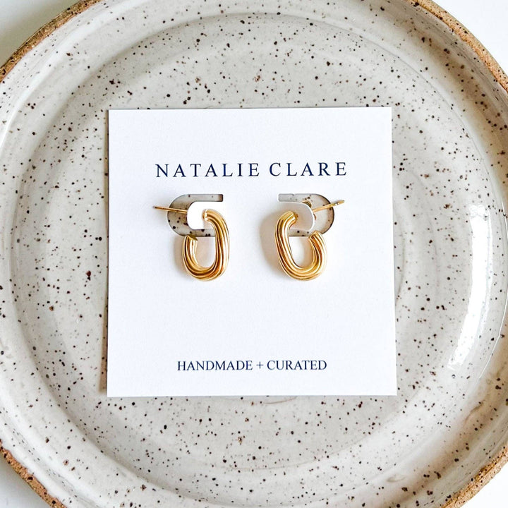 A pair of gold abstract loop hoop earrings displayed on a white Natalie Clare card, set against a speckled ceramic plate.