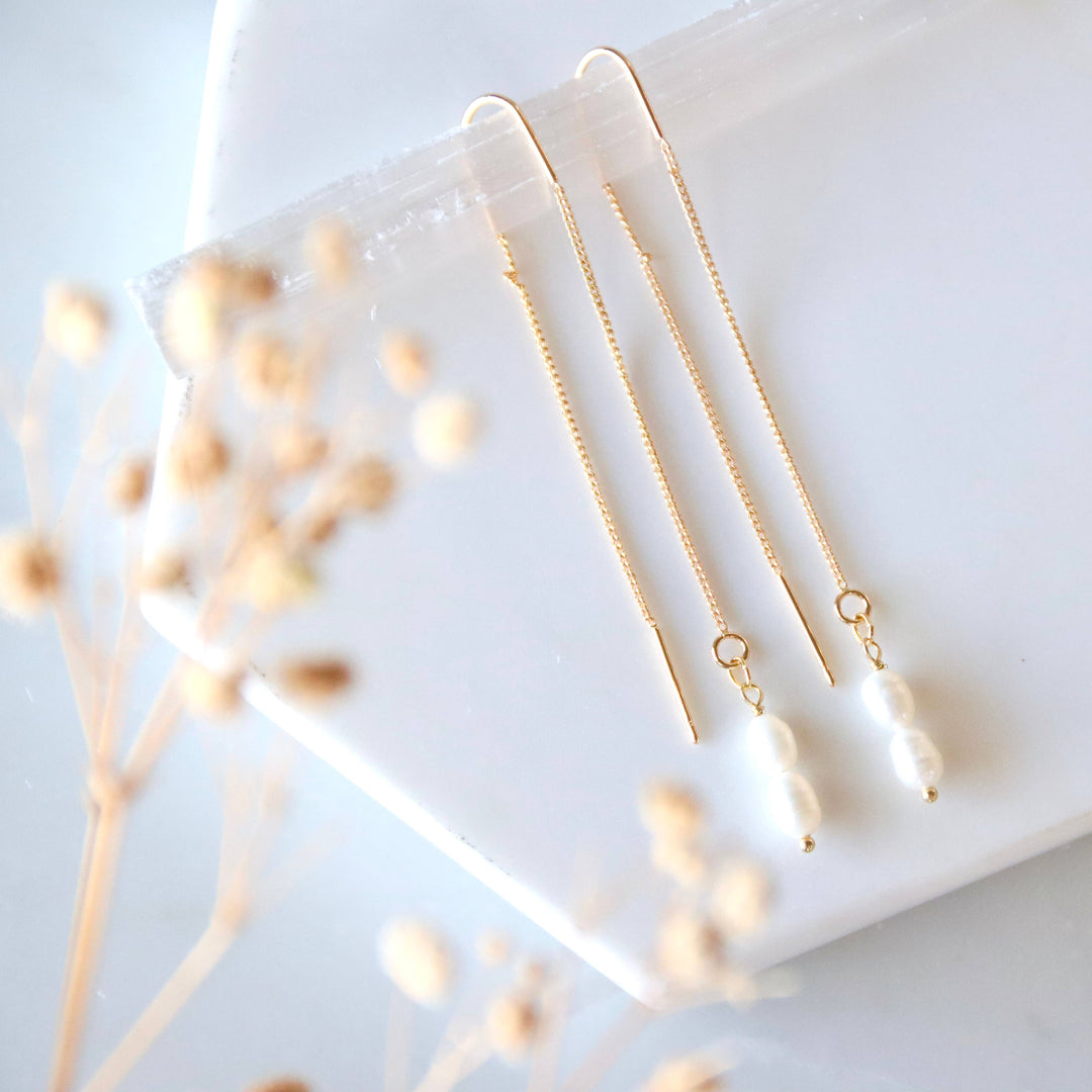 Gold and pearl dangling earrings on a white surface with dried flowers.
