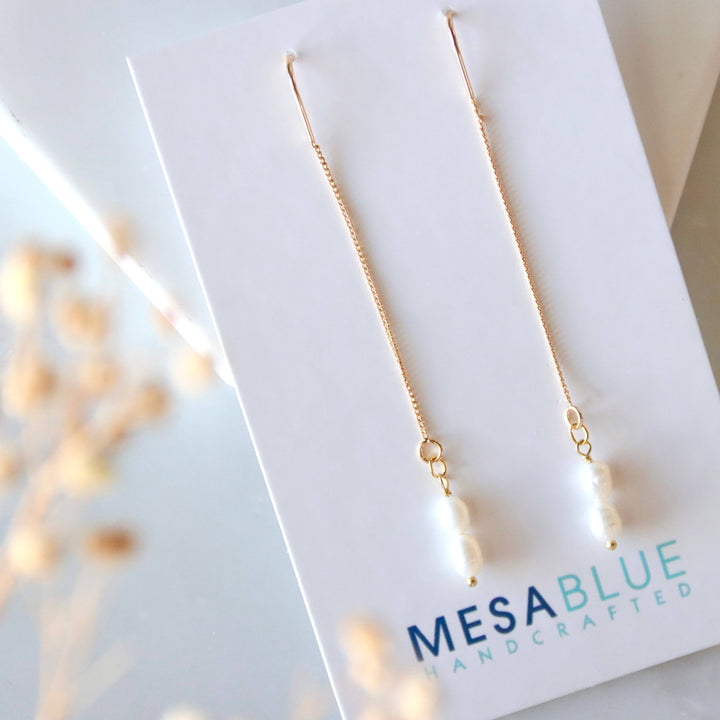 Gold and pearl dangling earrings on a Mesa Blue branded card with a blurred background