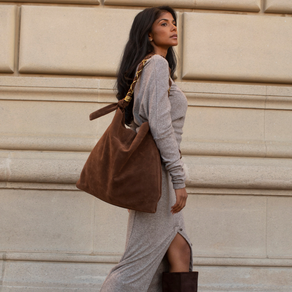 A woman in a gray knit dress and brown boots carries a large brown suede shoulder bag with a gold chain strap, standing against a beige stone wall.