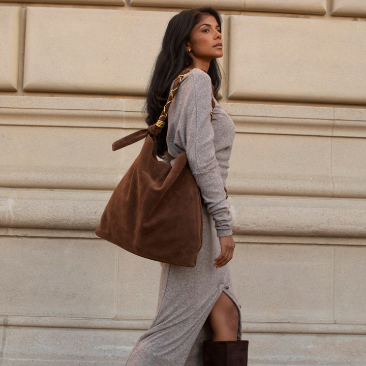 A woman in a gray knit dress and brown boots carries a large brown suede shoulder bag with a gold chain strap, standing against a beige stone wall.