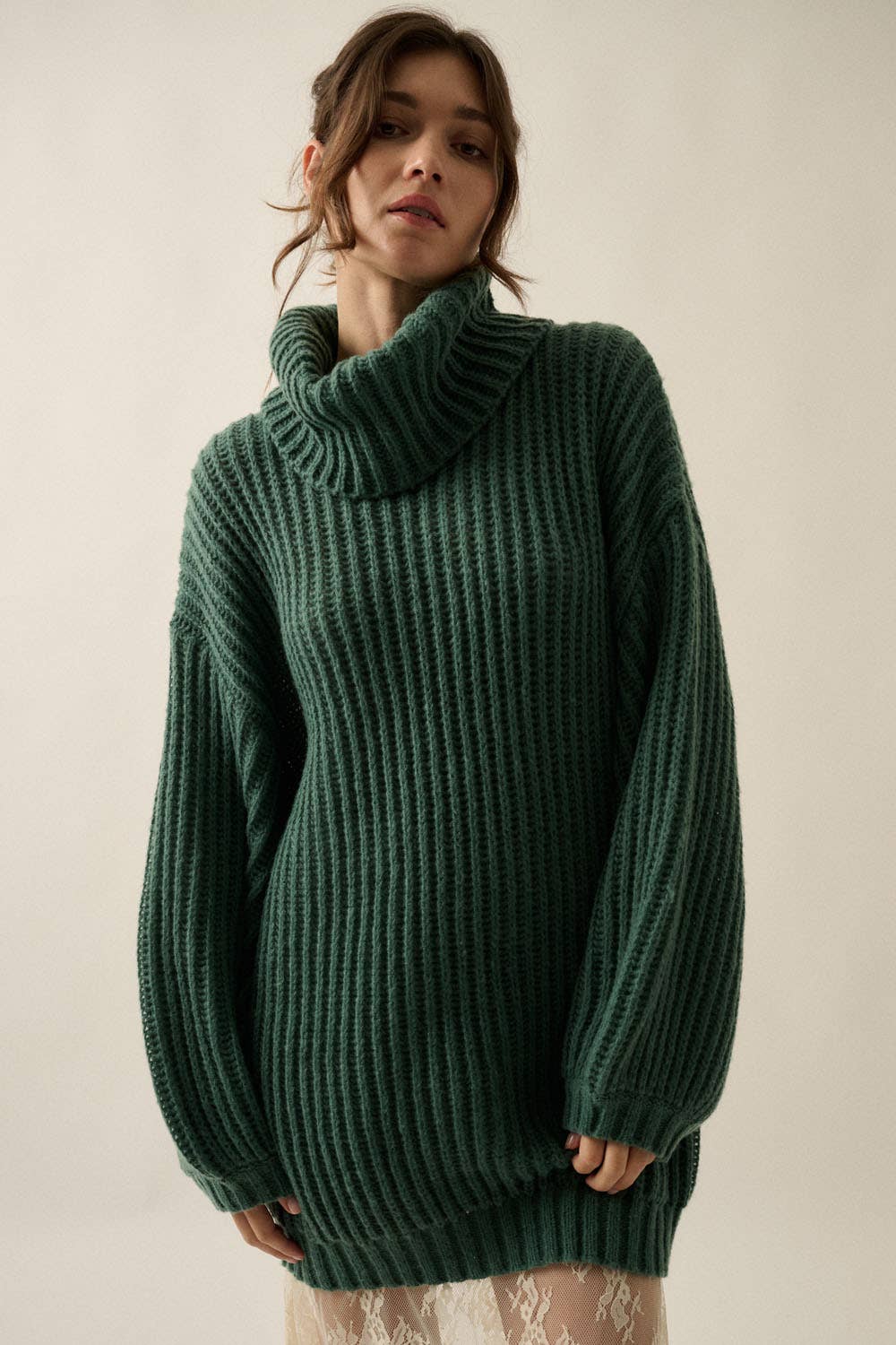 A woman wearing an oversized dark green ribbed knit turtleneck sweater, photographed against a plain light beige studio background.