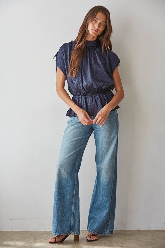 A woman modeling a dark blue animal print short-sleeve top with a peplum waist and wide-leg blue jeans, standing against a plain off-white wall.