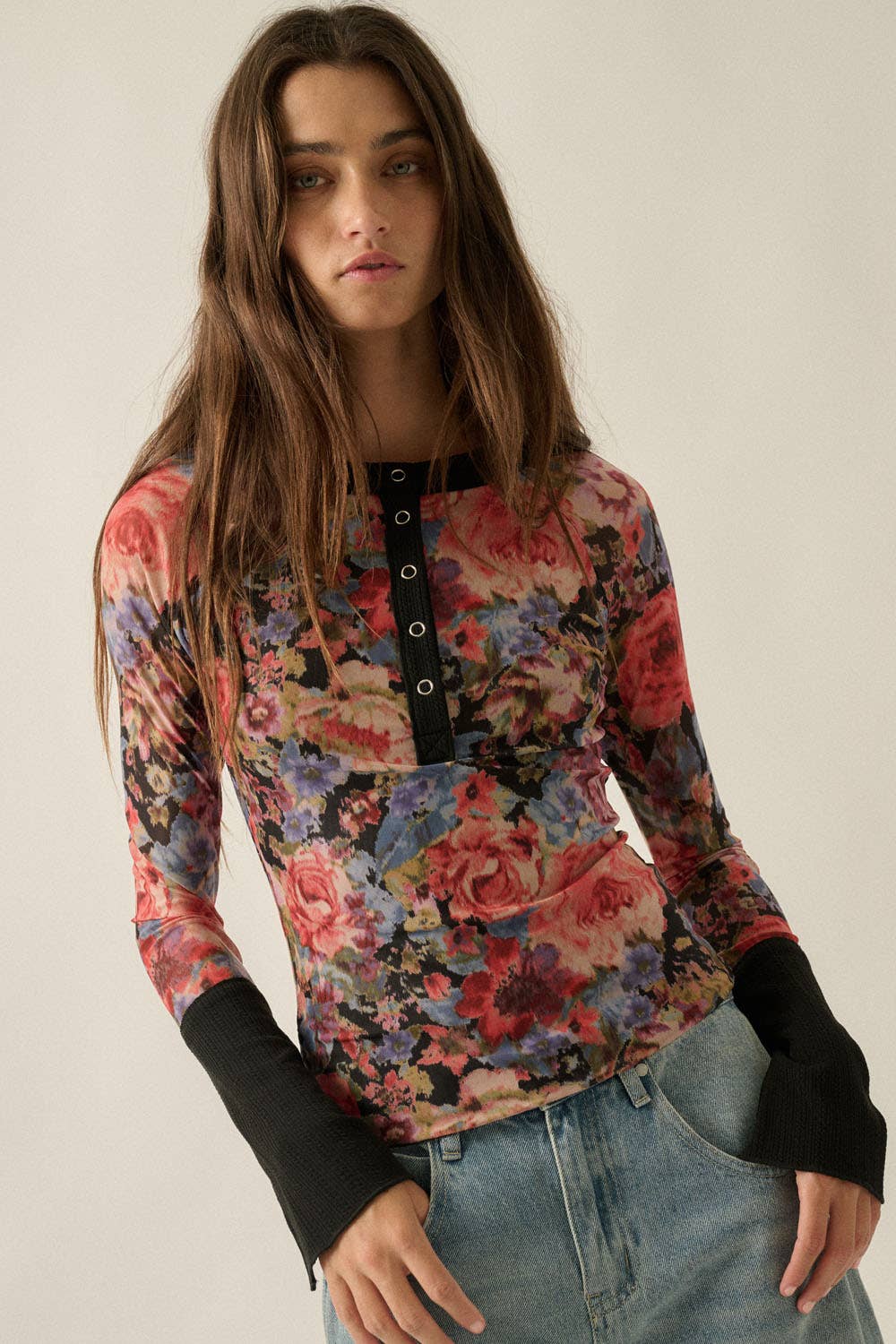 A woman wearing a long-sleeve floral henley top with black button placket and black ribbed cuffs, paired with light-wash blue jeans, against a plain light beige background.
