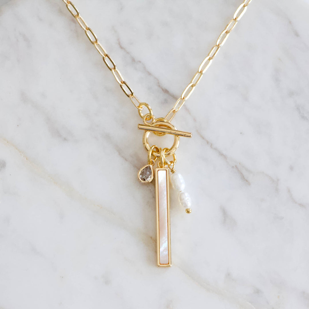 Gold necklace with a vertical bar pendant and mother of pearl charm on a marble background