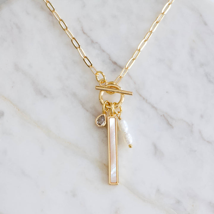 Gold necklace with a vertical bar pendant and mother of pearl charm on a marble background