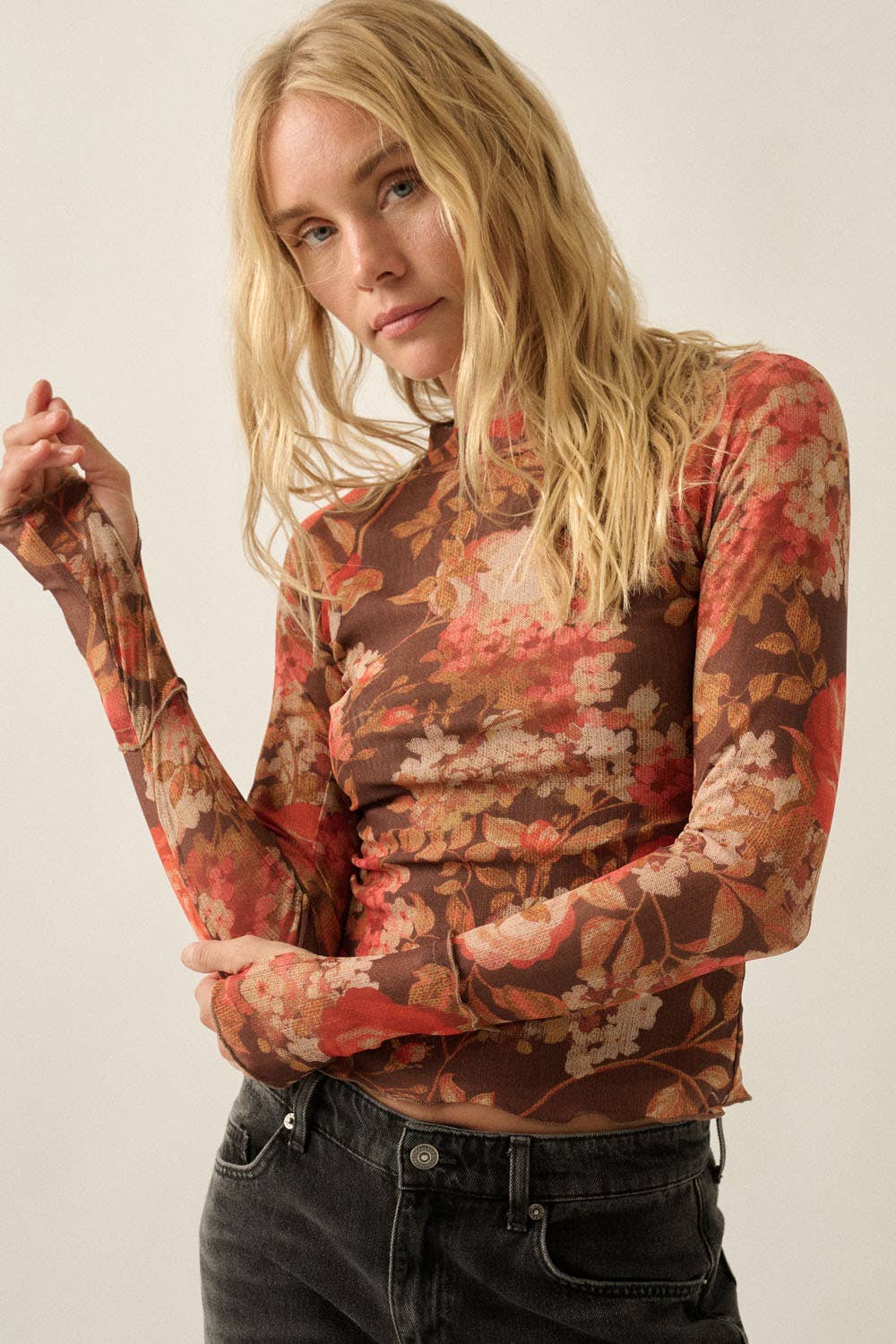 A woman wearing a long-sleeve mock-neck top with a brown, red, and beige floral pattern, paired with dark gray jeans, against a plain light background.