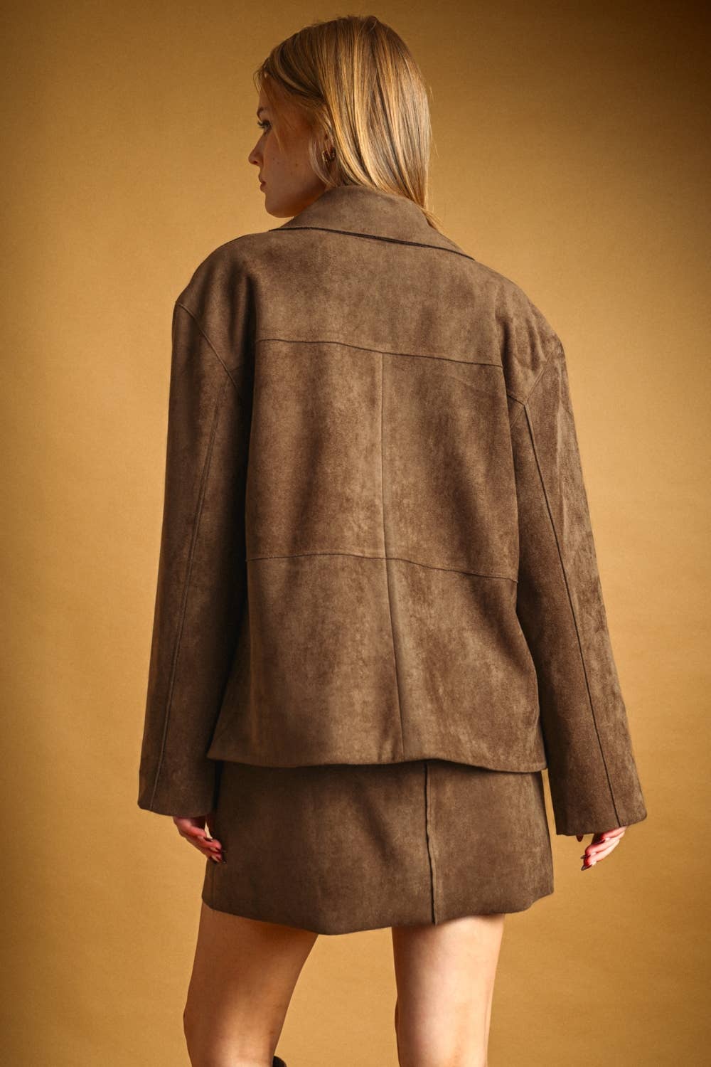 A woman shown from the back wearing a brown faux suede jacket and matching mini skirt against a warm tan studio background.