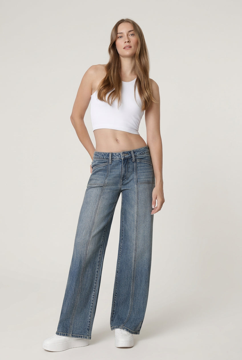 A woman modeling a pair of wide-leg blue jeans with a white crop top and white sneakers, standing against a clean white studio background.