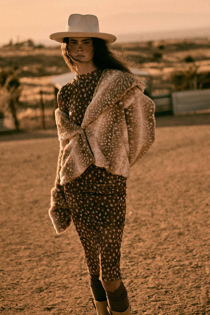 A woman wearing a brown and beige polka dot midi dress with a fuzzy beige sherpa jacket, a white cowboy hat, and dark boots, standing in a dusty outdoor desert landscape.
