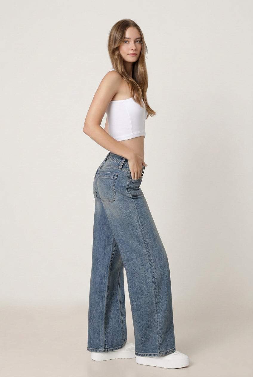 A woman modeling a pair of straight leg blue denim jeans with a white strapless crop top and white sneakers, photographed against a clean white studio background.