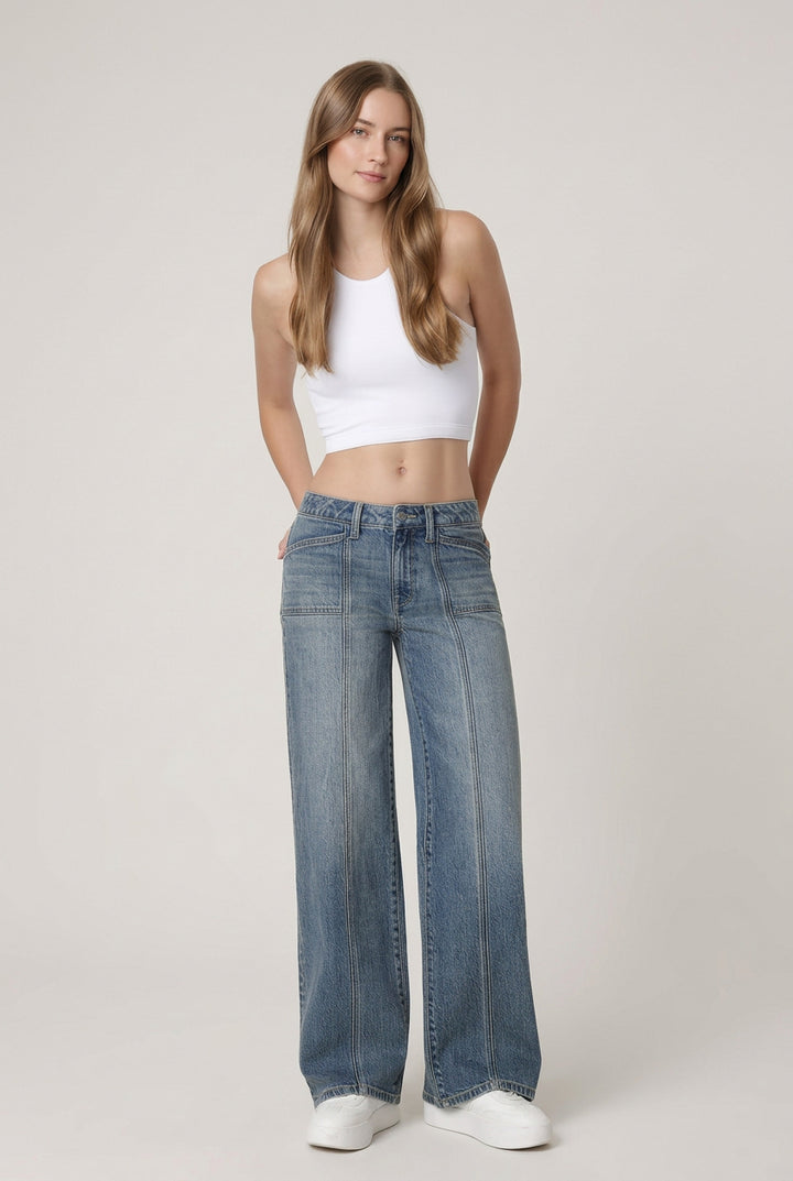 A woman modeling a pair of wide-leg blue jeans with a white crop top and white sneakers, standing against a plain white studio background.