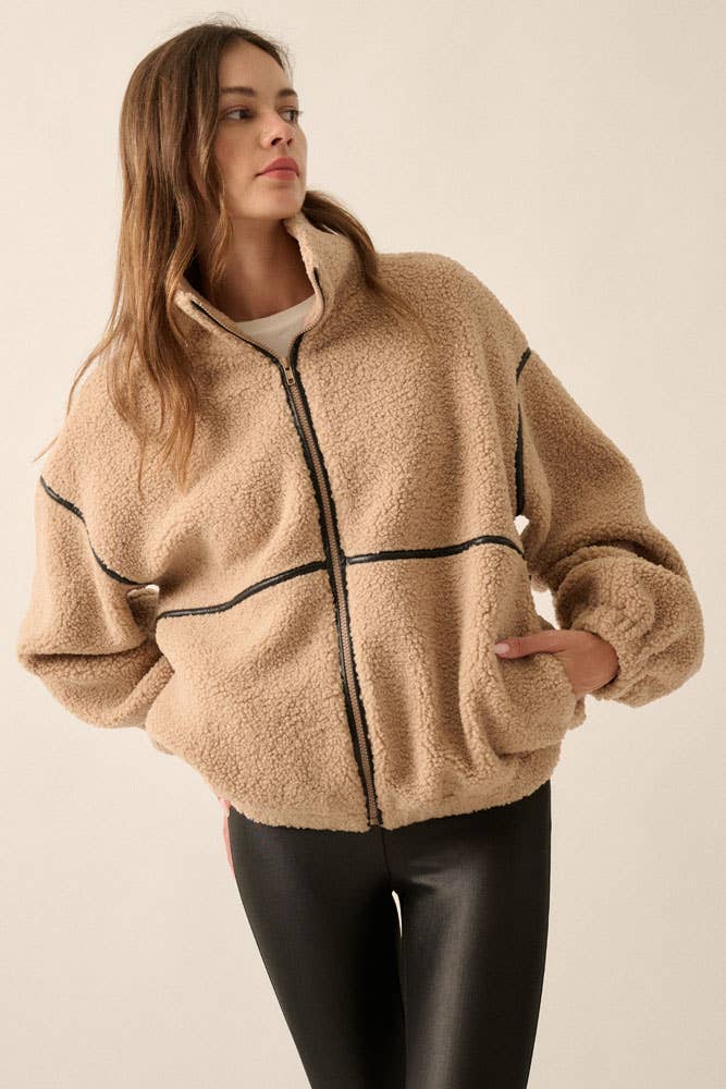 A woman wearing a beige sherpa fleece zip-up jacket with black faux leather contrast piping and black leather-look pants, photographed against a plain light beige studio background.