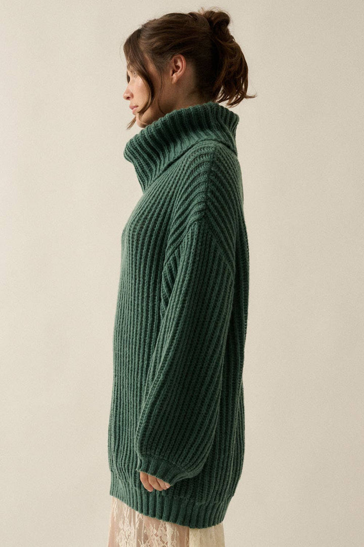 A woman shown in side profile wearing an oversized dark green ribbed knit turtleneck sweater against a plain light beige studio background.