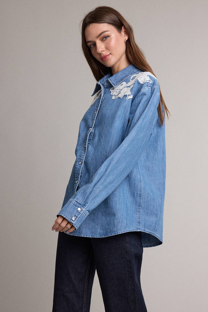 A woman wearing a light blue denim button-up shirt with white floral embroidery on the shoulders, paired with dark blue jeans, photographed against a solid light gray studio background.
