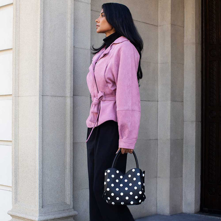 A woman in a pink belted jacket and black pants stands in profile against a stone building, holding a black tote bag with white polka dots.
