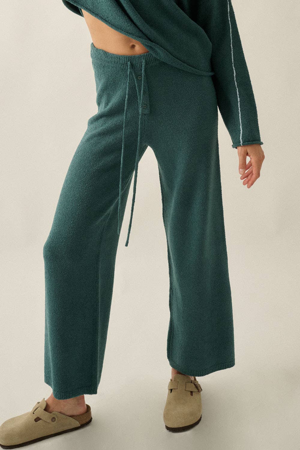 A person wearing dark teal knit wide-leg pants with a drawstring waist, paired with a matching knit top and beige slip-on shoes, photographed against a plain light background.