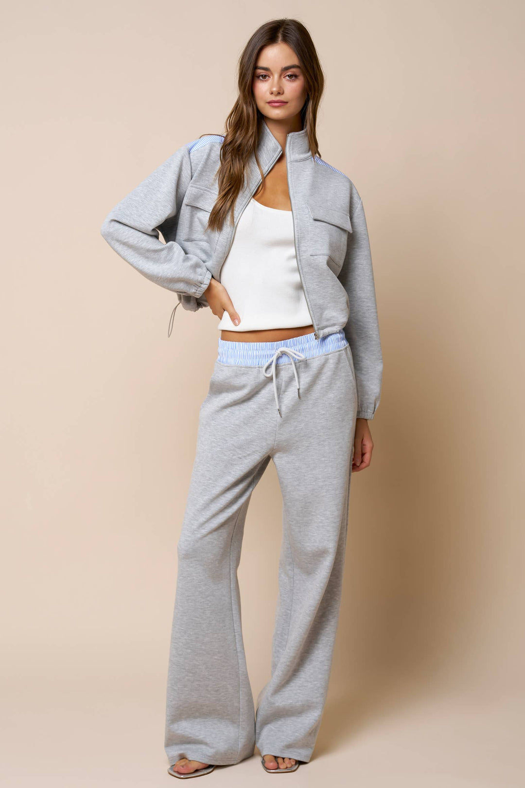 Woman wearing a light gray sweatshirt jacket and matching sweatpants with a white top against a beige background