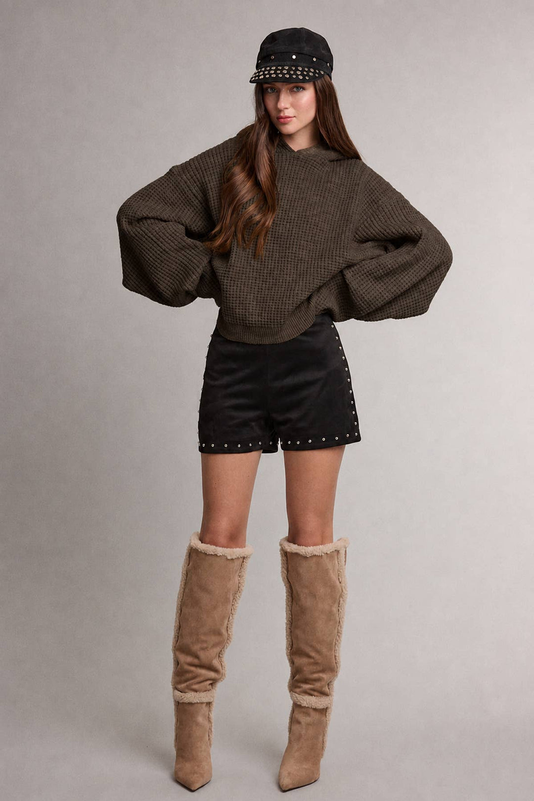 A model wearing a dark brown waffle-knit cropped sweater with hood, black studded shorts, a black studded cap, and tall tan suede boots with faux-fur trim, standing against a neutral gray studio background.