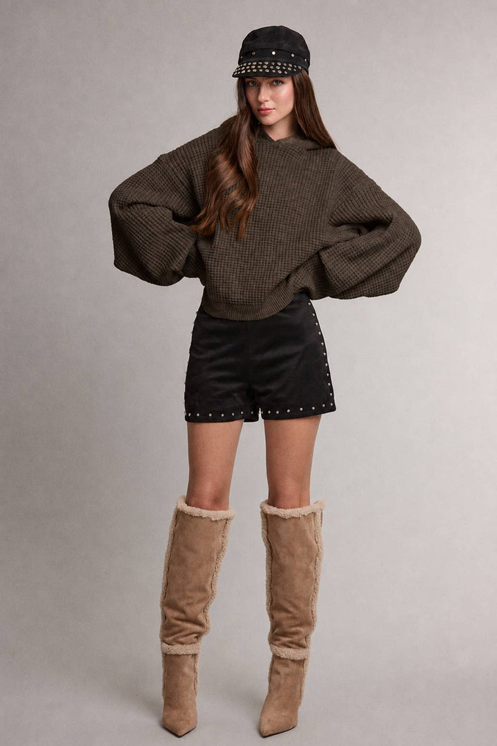 A model wearing a dark brown waffle-knit cropped sweater with hood, black studded shorts, a black studded cap, and tall tan suede boots with faux-fur trim, standing against a neutral gray studio background.