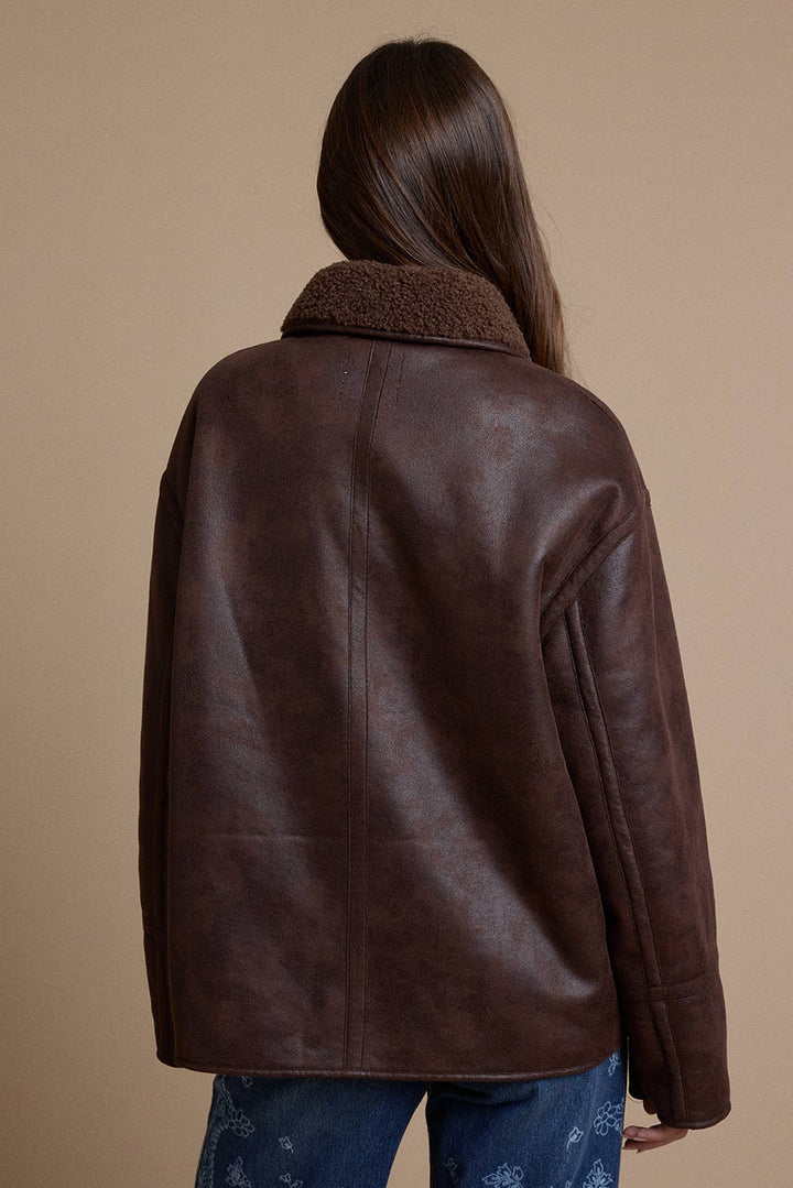 A woman viewed from behind wearing a dark brown faux-leather jacket with a plush brown sherpa collar, paired with blue jeans, against a solid beige background.