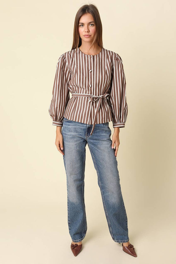 A woman modeling a brown and white vertically striped long-sleeve blouse with a tie waist, paired with blue jeans and brown pointed shoes, against a solid beige background.