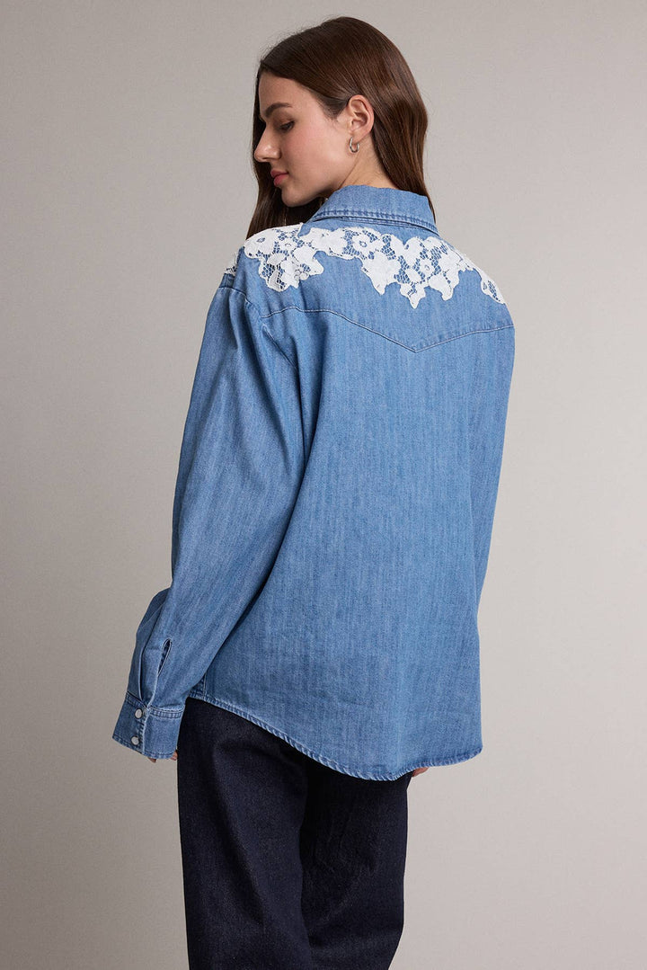 A woman wearing a light blue denim shirt with white floral lace embroidery across the shoulders, shown from a side-back angle against a plain light gray studio background.