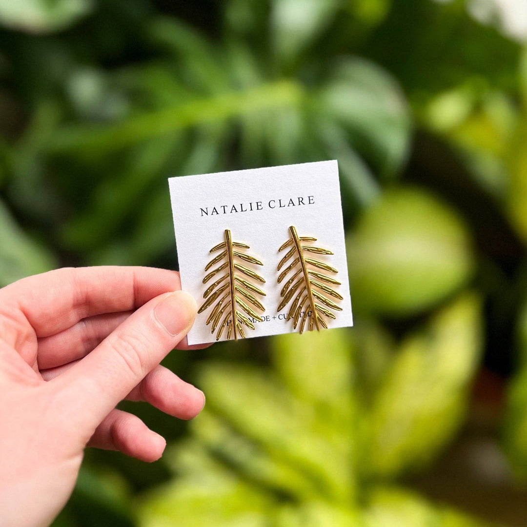 Hand holding a card with gold leaf earrings and 'Natalie Clare' branding against a green leafy background.