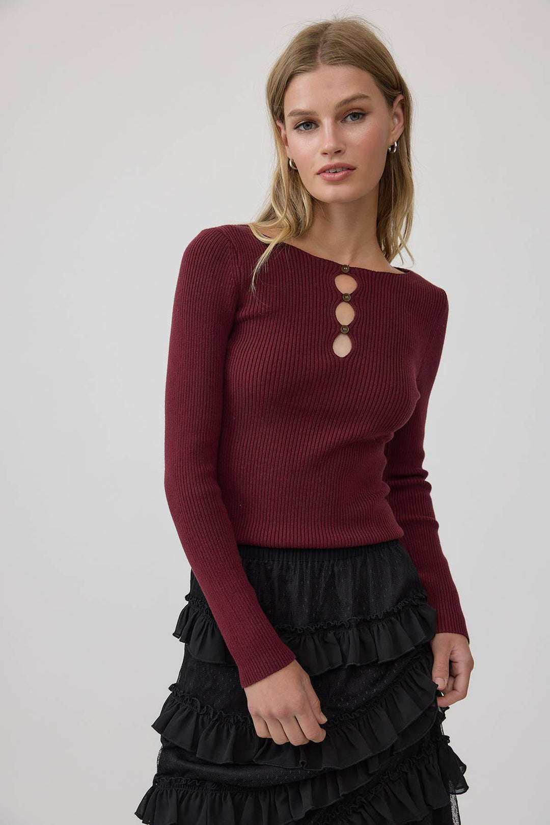 A woman modeling a ribbed long-sleeve knit top in deep burgundy with three large round cutout buttons at the neckline, paired with a black ruffled skirt, against a light gray studio background.