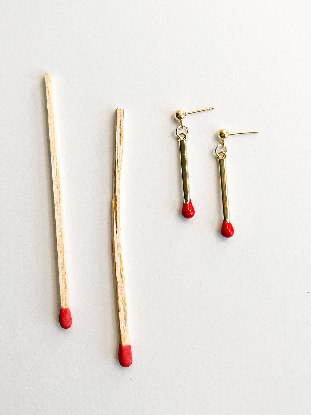 Gold drop earrings resembling matchsticks with red tips on a light gray background