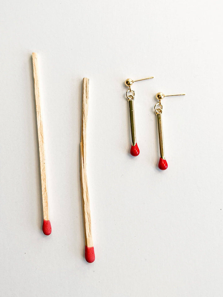 Gold drop earrings resembling matchsticks with red tips on a light gray background